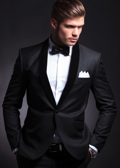 waist-up picture of an elegant young fashion man in tuxedo looking away from the camera while holding hands in pockets.on black background